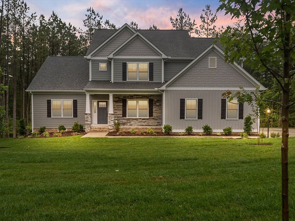Two-story gray house at Shirley Commons with front lights on, large lawn, shrubs, and trees at sunse
