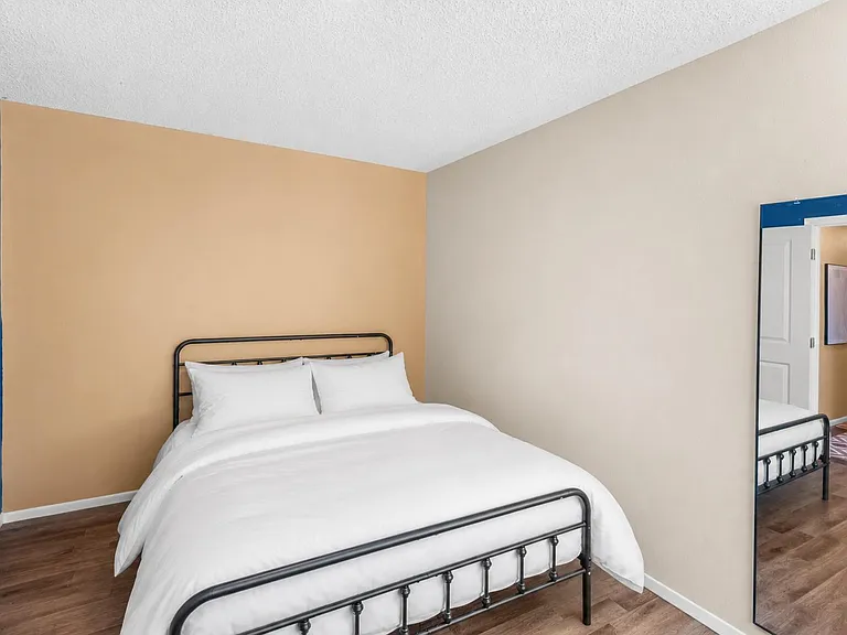 Furnished bedroom with a full-size bed, hardwood-style flooring, and a full-length mirror near the entry.