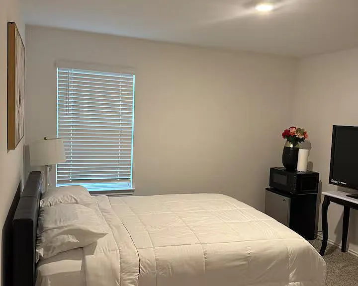 Private bedroom with a full-size bed, window with blinds, and carpeted floors, plus space for a small desk or TV setup.
