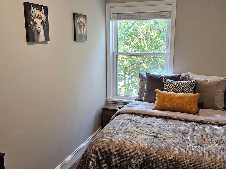 Furnished bedroom with a full bed, nightstand, work desk, mounted Smart TV, and a large window with blinds overlooking views.