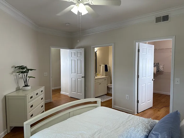 Bright bedroom featuring elegant crown molding, composite wood flooring, and a cozy bed. Adjacent walk-in closet and modern en-suite bathroom add luxury and convenience. Bright bedroom featuring elegant crown molding, composite wood flooring, and a cozy b