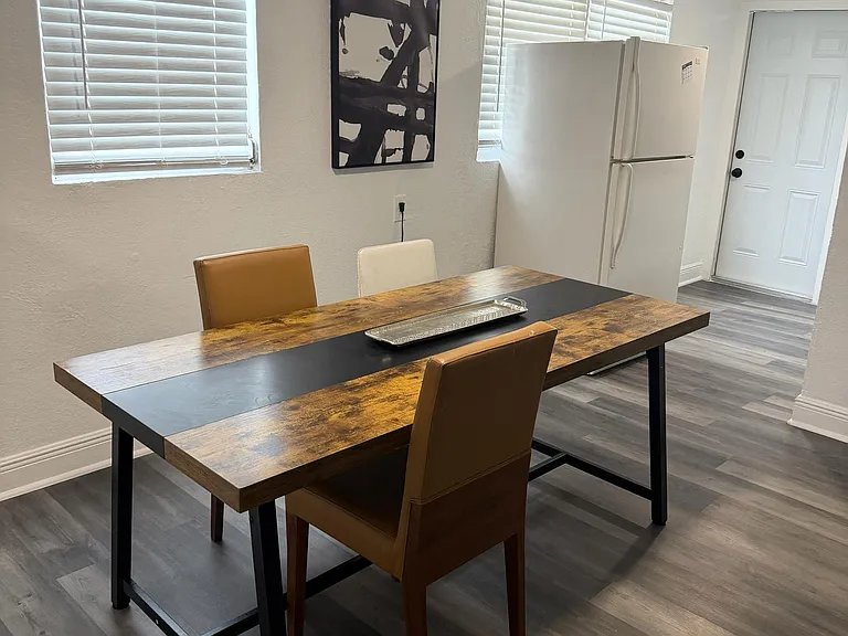 Dedicated dining area with a long table, seating for three, and nearby refrigerator, plus window blinds for light control.