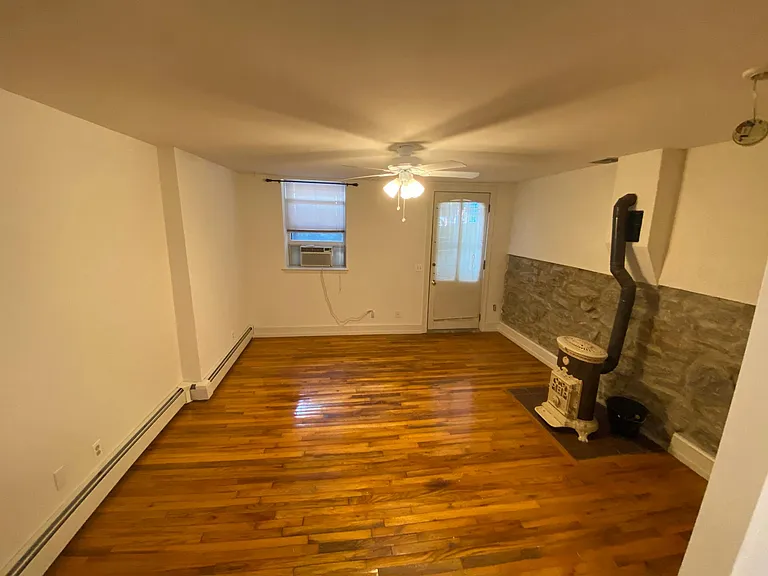 LIVING ROOM WITH HARDWOOD FLOOR.A FRANKLIN WORKING STOVE ON THE RIGHT. DOOR TO STREET LEVEL WINDOW WITH AIR CONDITIONING UNIT