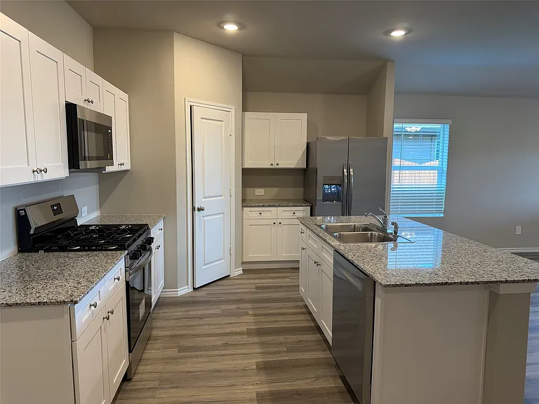 Kitchen featuring white cabinetry, stainless steel appliances, granite-style countertops, wood-finish flooring, and an island with a double basin sink
