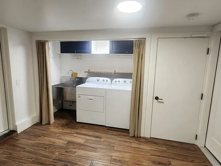 Downstairs, a flexible use utility room with laundry closet including new washer and dryer, an industrial utility sink, retractable drying racks, ceiling-mounted upper cabinets for storage, and curtains. Door access for garage, back yard & kitchen stairs