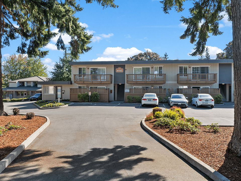 Front view of an apartment building with tan and gray siding, balconies, and resident parking in front.