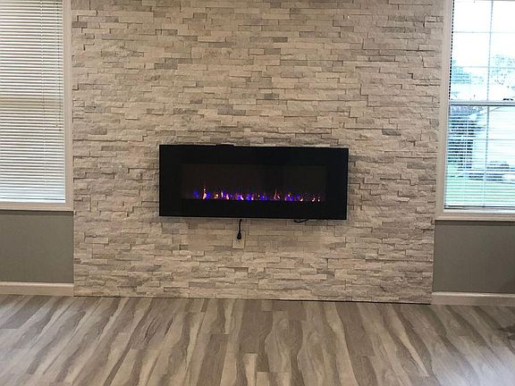 Enter into the foyer & to your left is a 18_ x 14_ Living Room with cathedral ceilings, custom window treatments, gray vinyl plank flooring, modern ceiling fan & floating fireplace with a custom white stone wall from floor to ceiling.