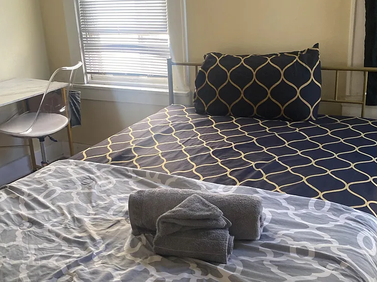 Furnished bedroom with a full bed, desk space, and a large window with blinds for natural light and privacy.