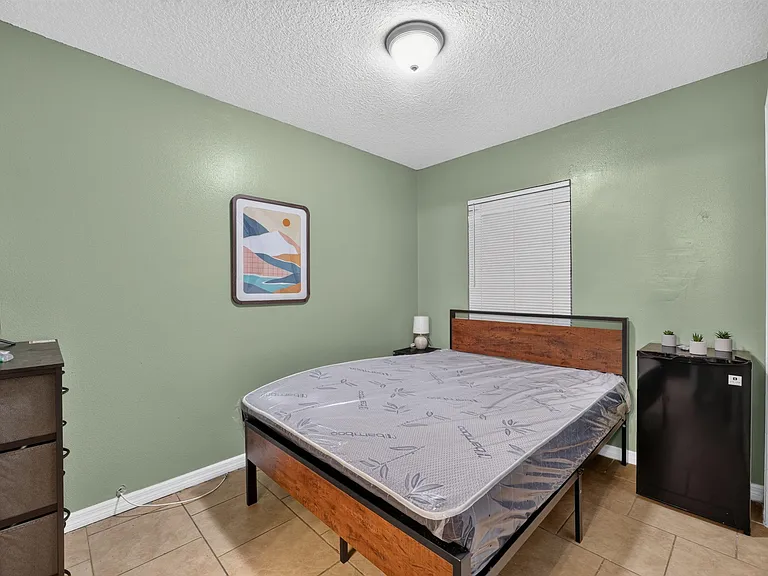 Furnished bedroom with a full-size bed, nightstand, mini fridge, and dresser, plus a window with blinds for privacy.