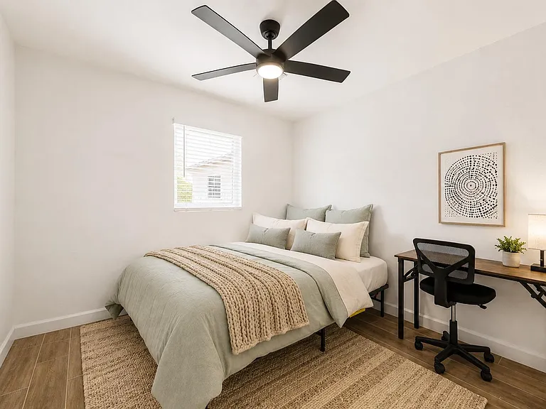 Furnished bedroom with a comfortable bed, ceiling fan, window with blinds, and a work desk for studying or remote work.