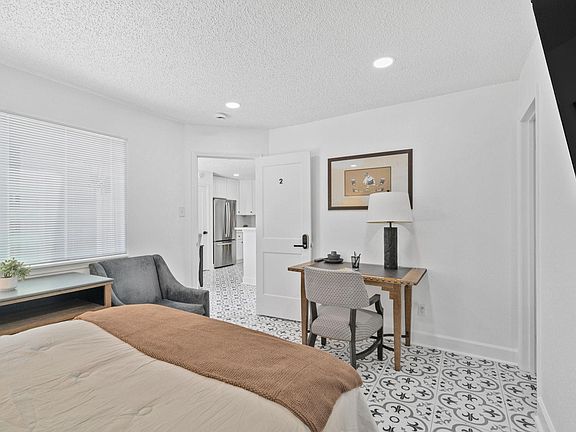 Furnished bedroom with patterned tile floors, a work desk, and window blinds, plus convenient access to the kitchen.