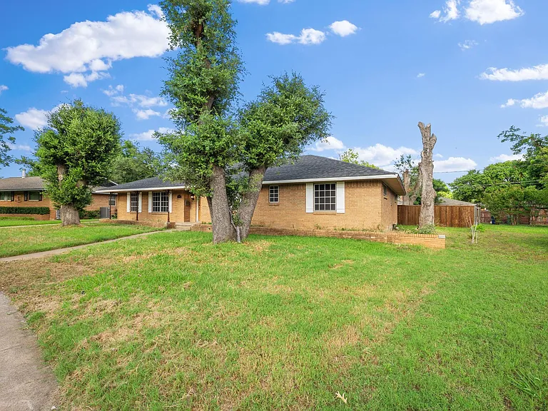 Single-story brick home with a spacious front lawn, mature trees, and a sidewalk leading to the covered entry.