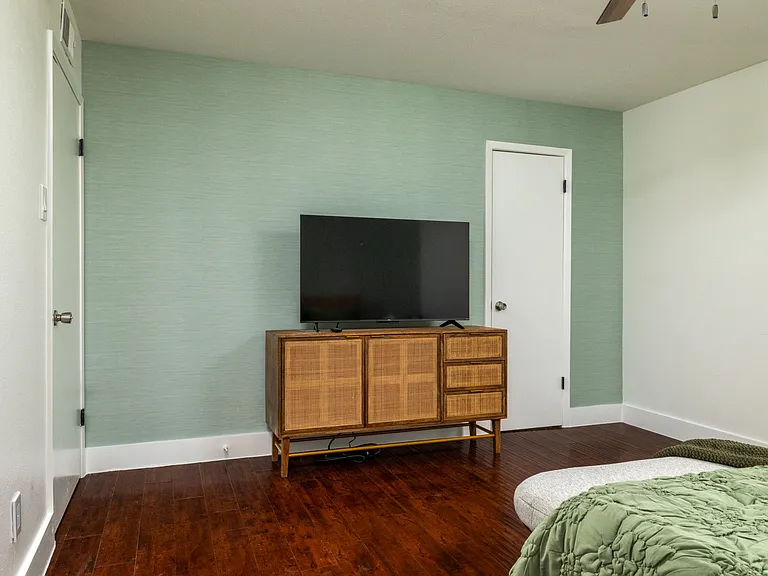 Rest in a calming bedroom with soft green tones, warm lighting, and a cozy bench at the foot of the bed. Enjoy a large TV, stylish dresser, and easy access to a spacious walk-in closet for a relaxing, comfortable stay.