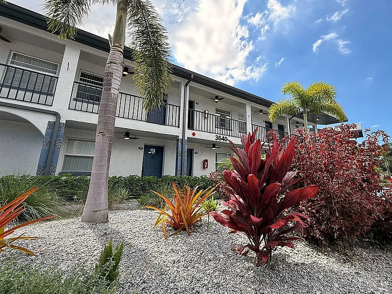 Charming two-story apartment building with modern balconies, surrounded by lush tropical landscaping and vibrant greenery under a bright blue sky.
