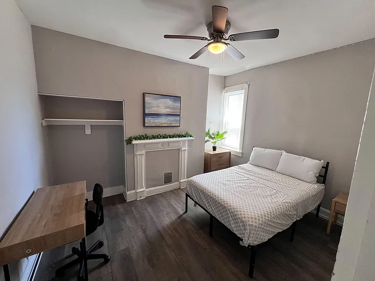 Furnished bedroom with a full bed, desk workspace, ceiling fan, and window light, plus decorative fireplace detail.