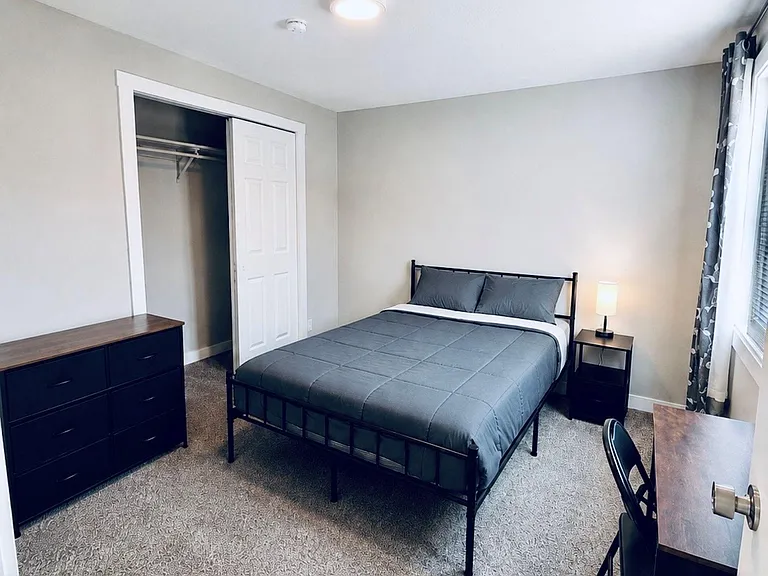Furnished bedroom with queen bed, dresser, desk workspace, and sliding-door closet, plus a large window with curtains.
