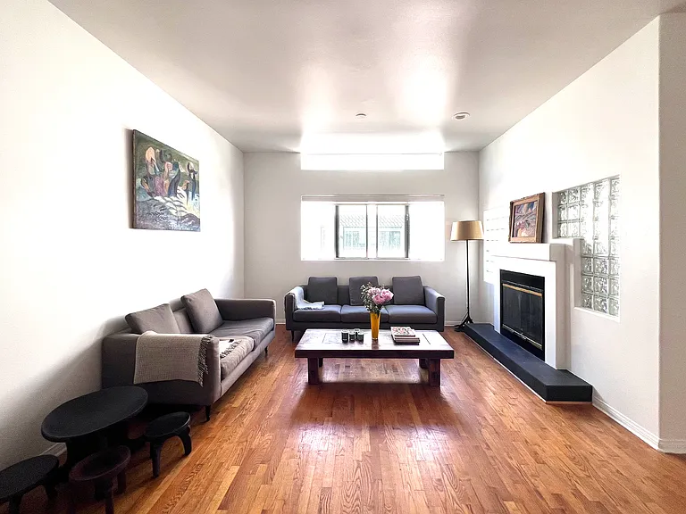 Bright open-concept living space with real wood floors, a working gas fireplace, a skylight, and large windows with roller shades, accented by curated artistic furniture.