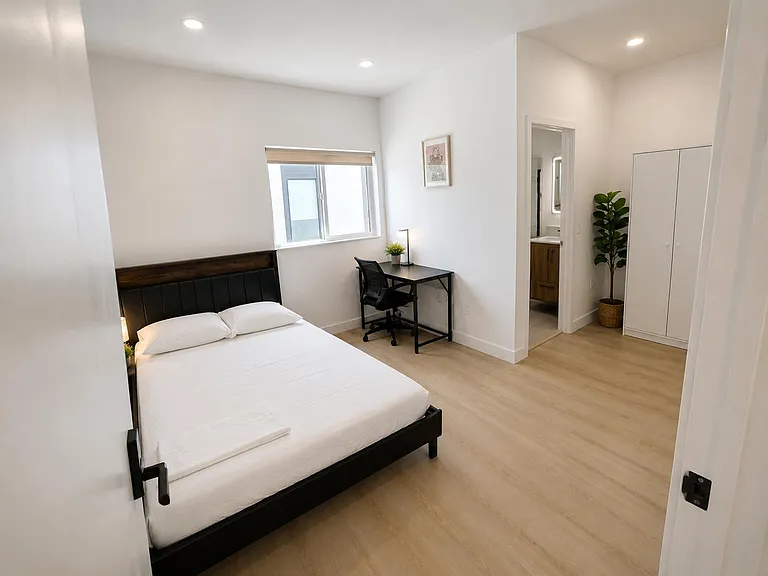Spacious furnished bedroom with a full bed, dedicated desk workspace, closet storage, and an adjacent bathroom access.