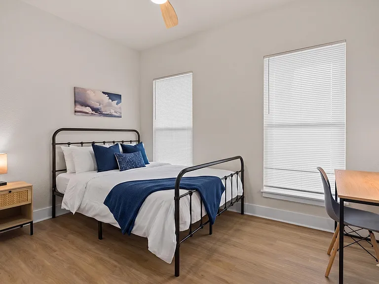Well-appointed bedroom with a full bed, large windows with blinds, and a dedicated desk workspace for studying or WFH.