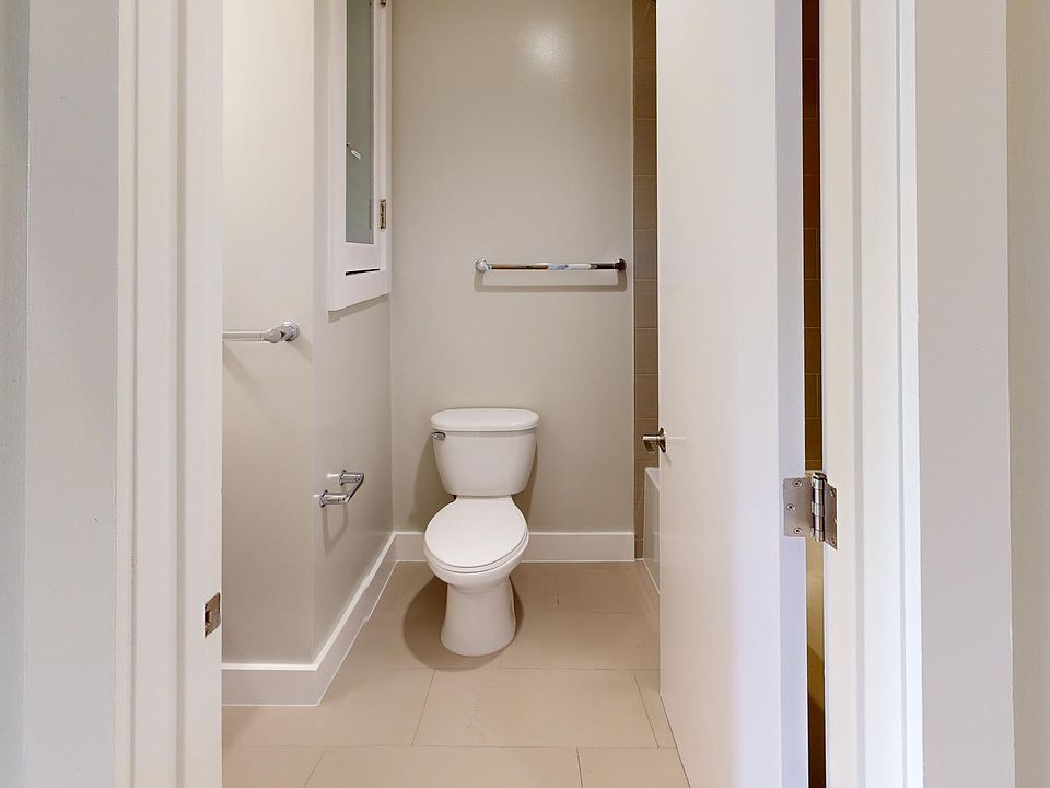 A small, bright bathroom with a toilet, walls, and tiled floor. A towel hangs on the wall. The open door adds a spacious feel.