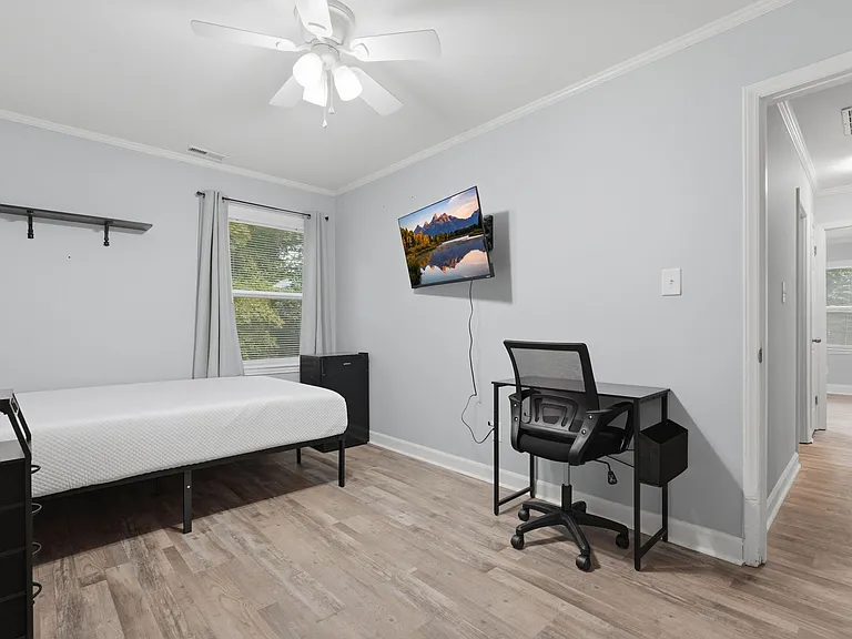Furnished bedroom featuring a bed, work desk, wall-mounted TV, and a window with curtains for natural light.