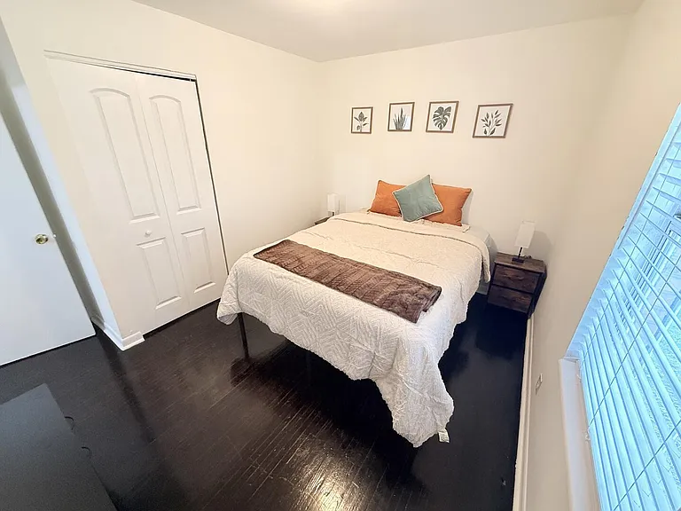 Private bedroom with dark wood floors, a full bed, bedside tables with lamps, and a large window with blinds.
