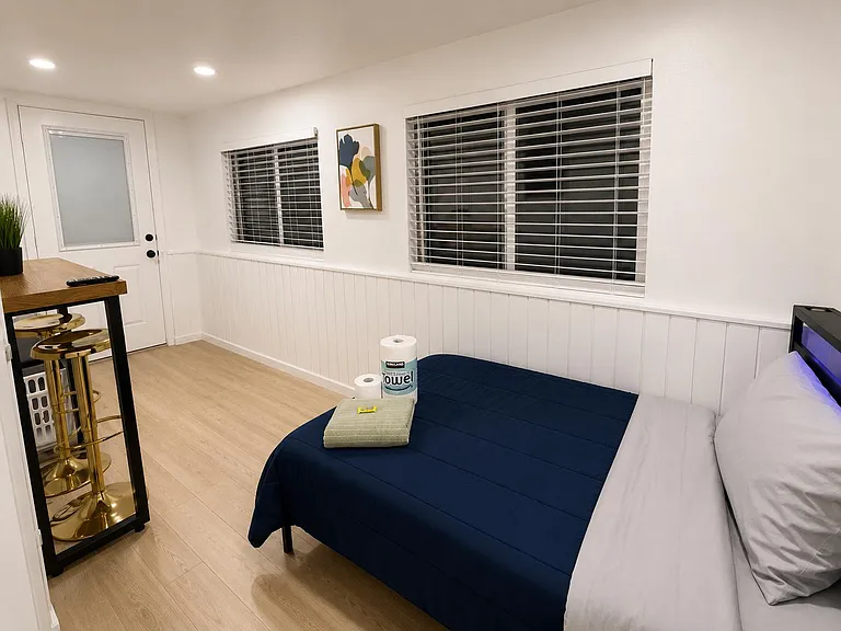 Furnished bedroom with a full bed, wood-style flooring, and windows with blinds; includes a small desk/bar area.