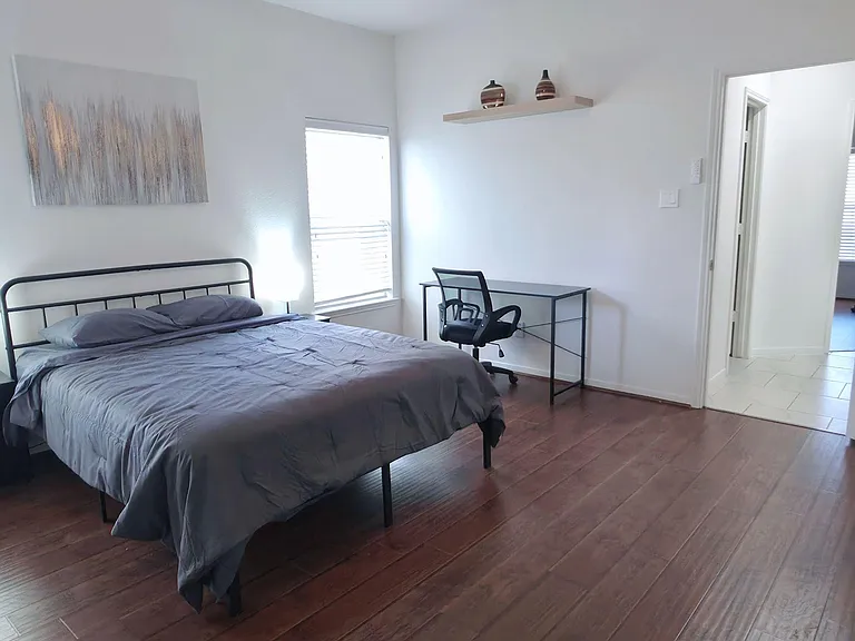 Furnished bedroom with wood-look floors, ceiling fan, window with blinds, and a dedicated desk/work area.