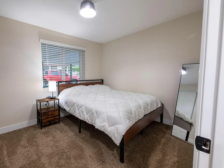 Comfortable bedroom with a full bed, nightstand lighting, carpeting, and a large window with blinds for privacy.