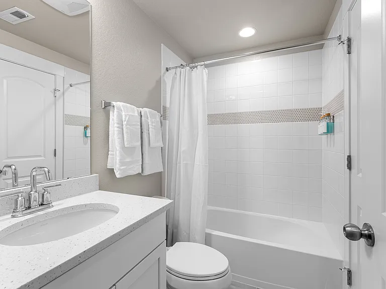 Bright bathroom with white vanity and quartz countertop. Features a clean bathtub with white tiles and a decorative accent strip, a shower curtain, and fresh towels. The space is well-lit and inviting for guests.