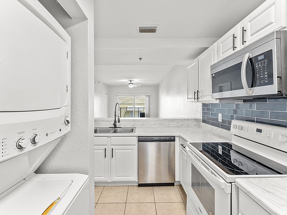 Modern kitchen with sleek white cabinetry, marble countertops, and stainless steel appliances, including a built-in microwave and stacked washer-dryer unit, leading to a bright living area.