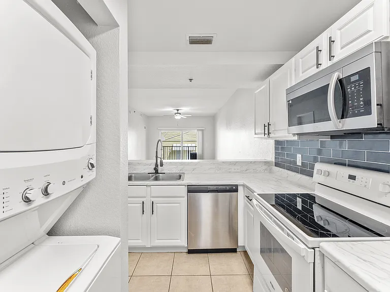 Modern kitchen with sleek white cabinetry, marble countertops, and stainless steel appliances, including a built-in microwave and stacked washer-dryer unit, leading to a bright living area.