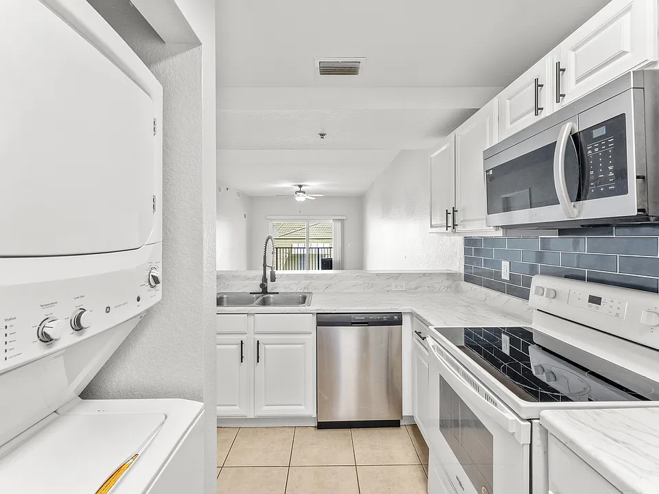 Modern kitchen with sleek white cabinetry, marble countertops, and stainless steel appliances, including a built-in microwave and stacked washer-dryer unit, leading to a bright living area.
