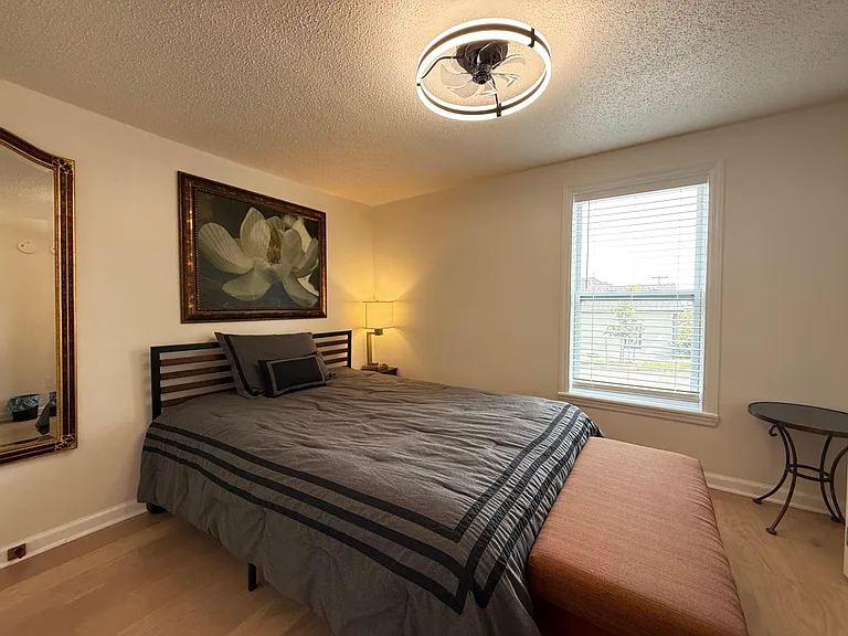 Furnished bedroom featuring a comfortable bed, bedside Ottoman with lamp, and a large window with blinds for natural light.