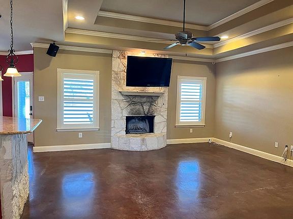 This spacious living room is perfect for entertaining. It has a tray ceiling, stone wood-burning fireplace, and stained concrete floors. The wall-mounted TV, surround sound system and ceiling fan add to the ambiance, and the plantation shutters add a grea