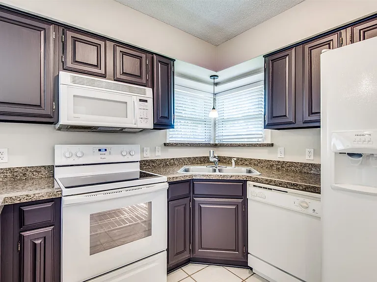 Kitchen featuring dark wood cabinetry, white appliances, speckled countertops, a corner sink with a pendant light, and tiled flooring