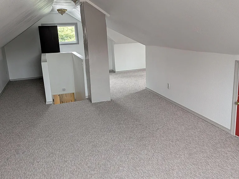 Carpeted upper-level room with sloped ceilings, overhead lighting, and a window for natural light.