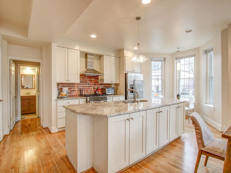 Close up of kitchen with room for a small table in front of the window, lots of cabinets, and industrial exposed brick backsplash.