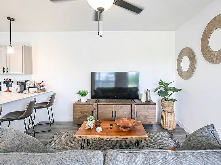 Modern living room with stylish decor, featuring a cozy gray sofa, elegant wood accents, and a sleek TV setup. Open-concept kitchen with chic bar stools and pendant lighting.