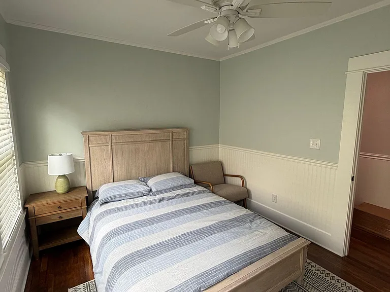 Furnished bedroom with a full bed, nightstand, and seating area, plus a ceiling fan and window with blinds.