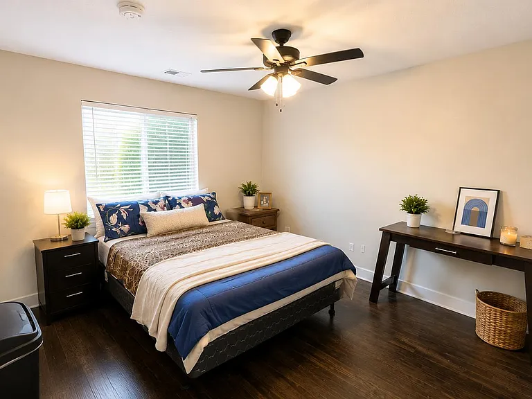 Spacious furnished bedroom with a full bed, nightstand, and dedicated desk area, plus a large window and ceiling fan.