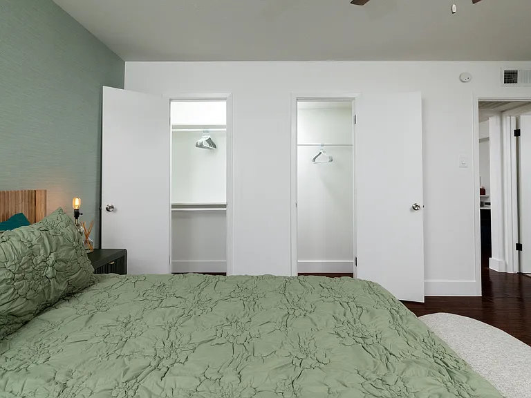 Rest in a calming bedroom with soft green tones, warm lighting, and a cozy bench at the foot of the bed. Enjoy a large TV, stylish dresser, and easy access to a spacious walk-in closet for a relaxing, comfortable stay.