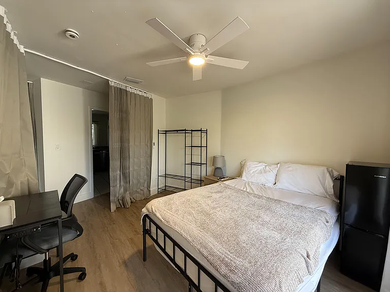 Furnished bedroom with ceiling fan, work desk, open shelving, and a mini fridge for convenient in-room storage.