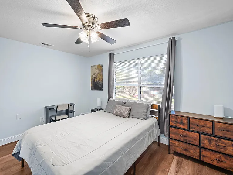 Well-sized bedroom with a large window, ceiling fan light, and space for a desk plus dresser storage.