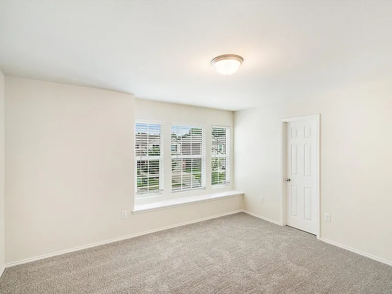 Spacious room featuring three large windows with blinds, a window seat, light cream walls, and gray carpet flooring