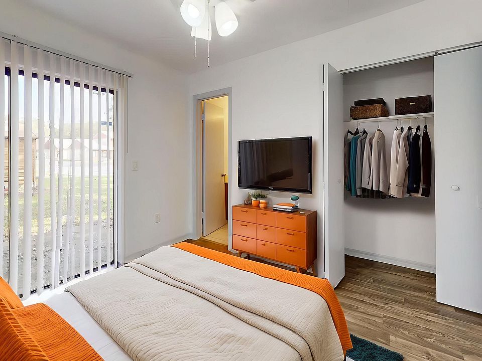 Chic bedroom with vibrant orange accents, featuring a cozy bed, sleek TV, and spacious closet, bathed in natural light.