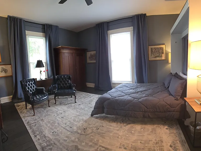 Panoramic view of second (front) bedroom from hallway entrance. Original fire place; Kentucky artwork featured (Alan Brewer and Joseph Petro); sitting area and vintage wardrobe for ample storage. Black out curtains; queen sized bed; ceiling fan.