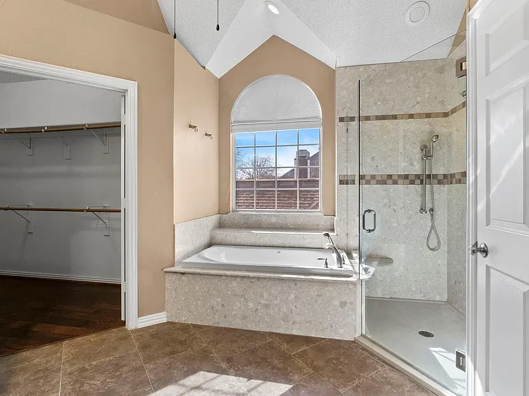 Spacious bathroom featuring a walk-in shower with tile surround, a built-in soaking tub with tiled steps, an arched window, and direct access to a walk-in closet with built-in shelving
