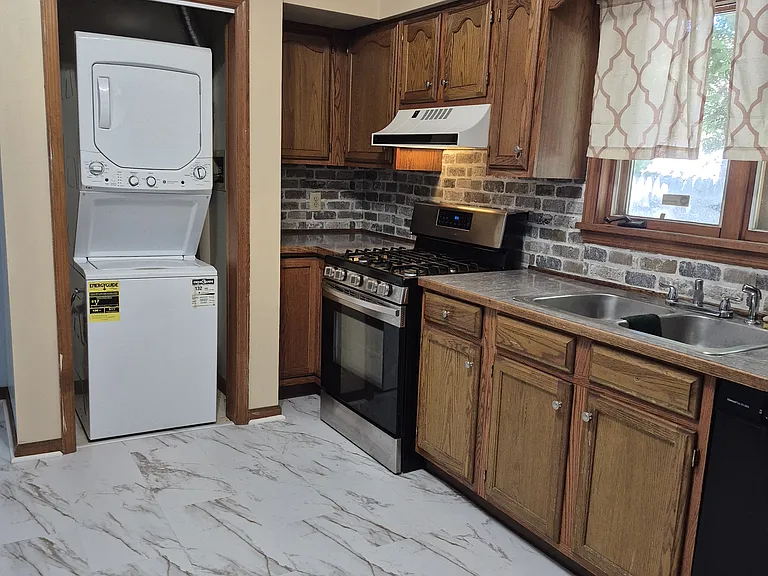 Kitchen view from Dining Room, new marble-look laminate tile flooring, stacked washer & dryer, gas stove, double stainless steel sink visible. Back door is just to left of washer, back room off to left also.