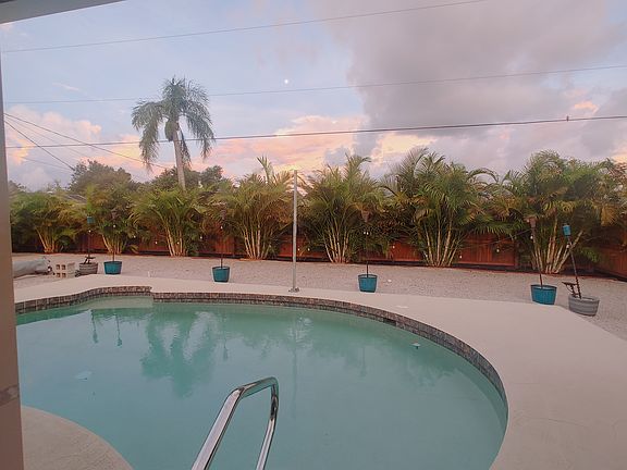 A tranquil backyard oasis complete with privacy landscaping, lush palms, and a refreshing pool set against a beautiful orangey-pink sunset sky.